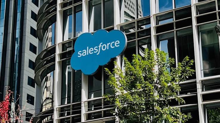 The blue Salesforce cloud logo on the glass front of a Salesforce office building, with a tree in the foreground