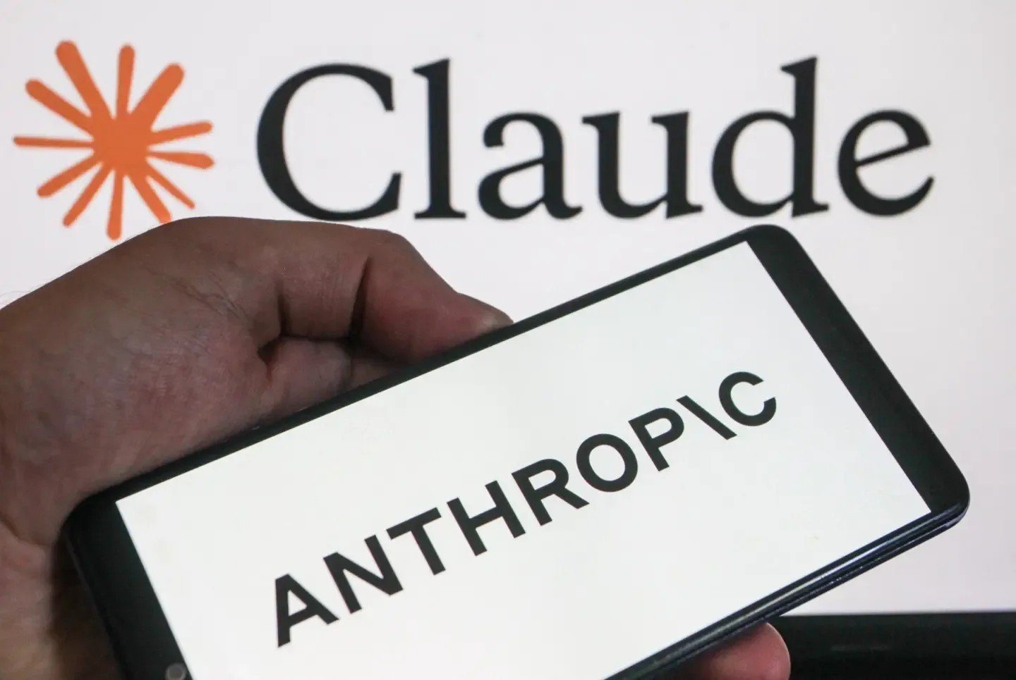 A hand holding a phone showing the Anthropic logo in front of a screen with the Claude logo