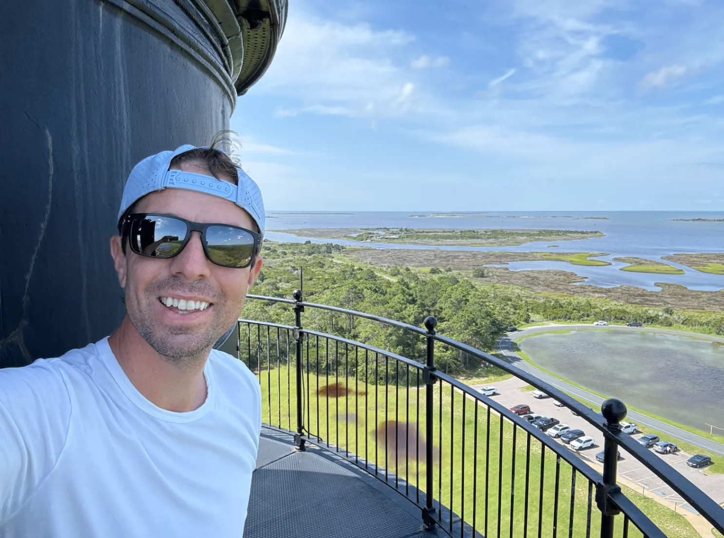 Keith Brown in sunglasses and a white T-shirt smiling on the black iron gallery at the top of a lighthouse, with marshland, a pond, a parking lot and open water far below