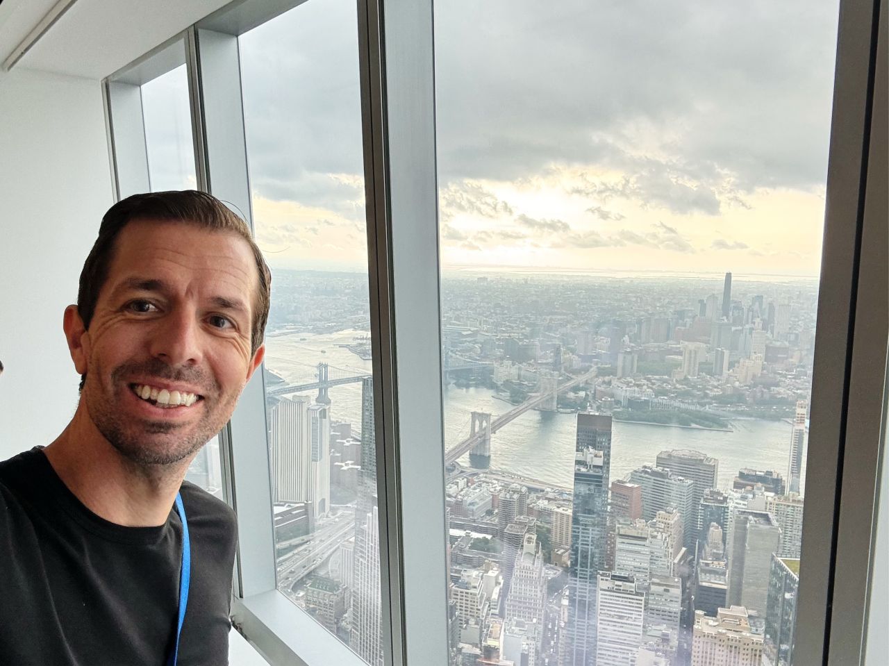 Keith Brown smiling at the window of One World Observatory, with the Brooklyn Bridge and the East River far below