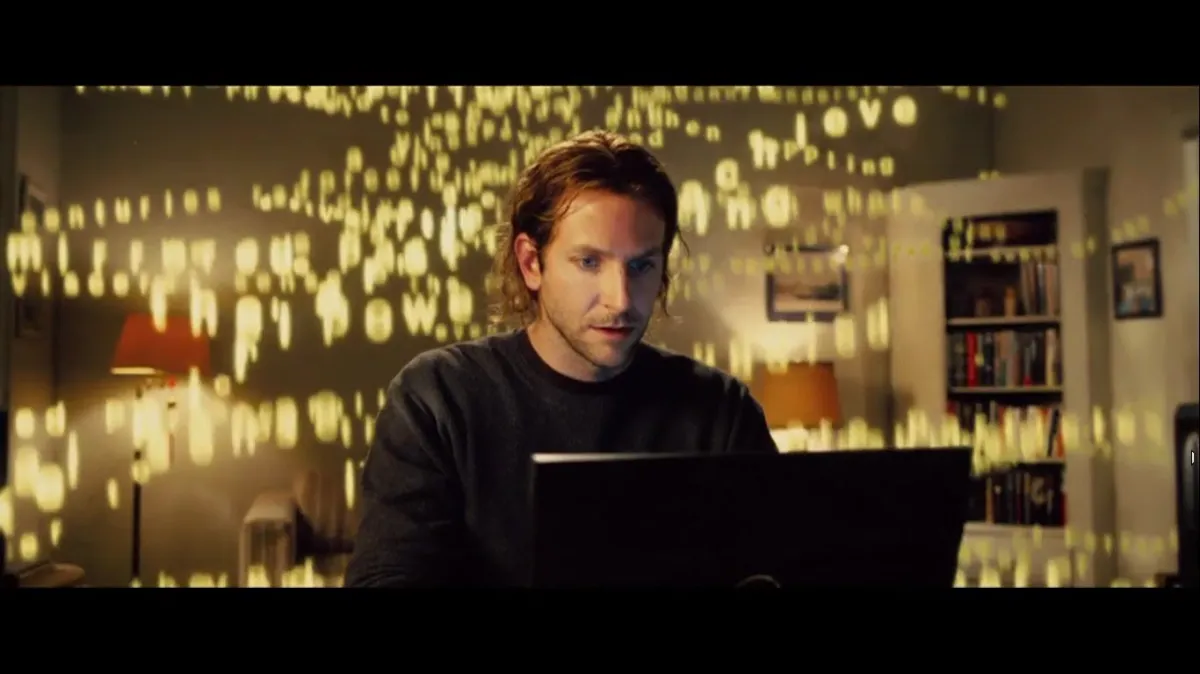Bradley Cooper as Eddie Morra typing on a laptop with glowing letters filling the room in the film Limitless