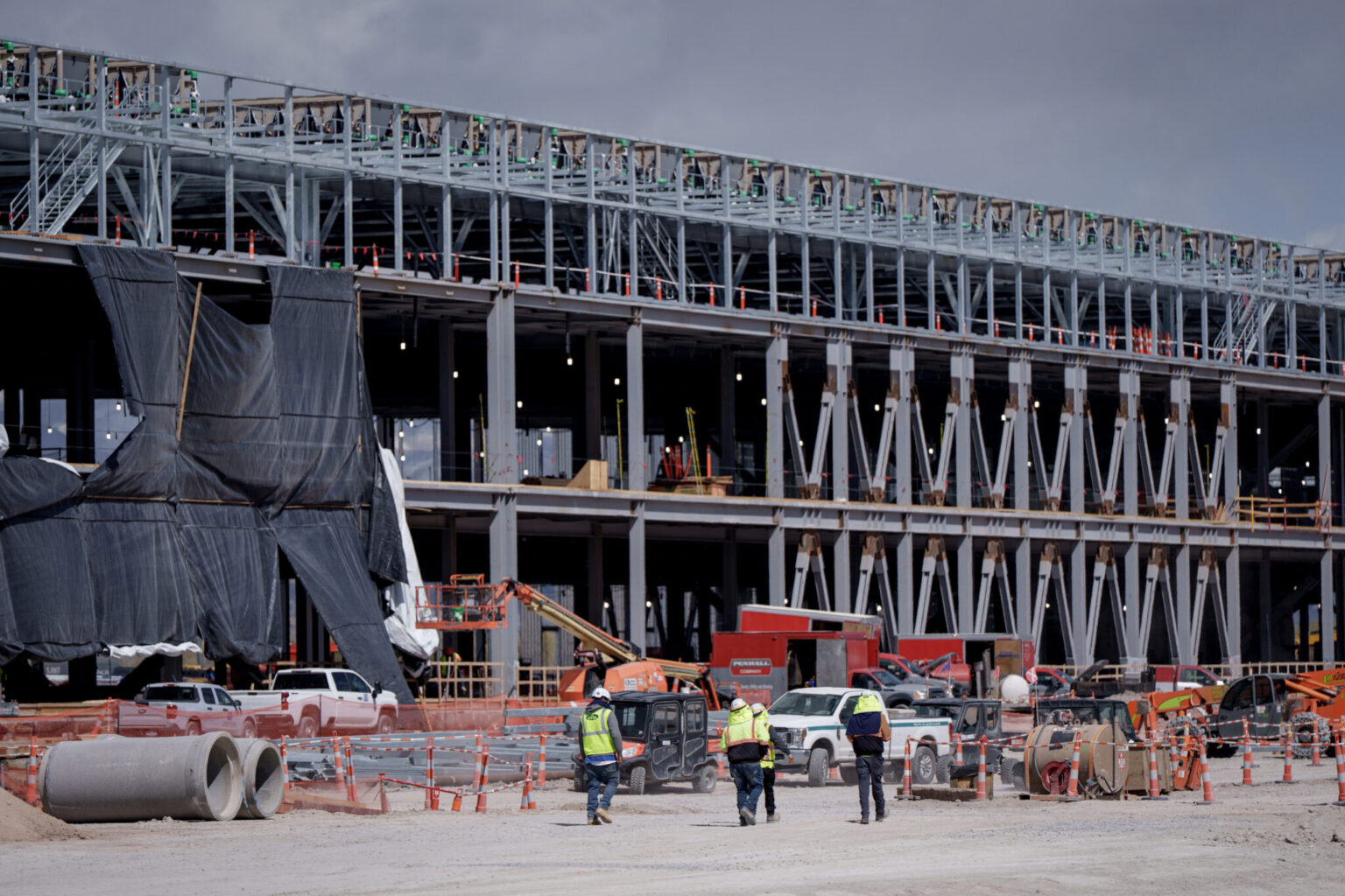 Steel frame of a large data center under construction, with workers in safety vests and equipment in the foreground
