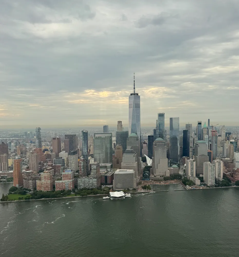 Aerial view of Lower Manhattan from over the Hudson River, with One World Trade Center rising above the skyline under grey clouds