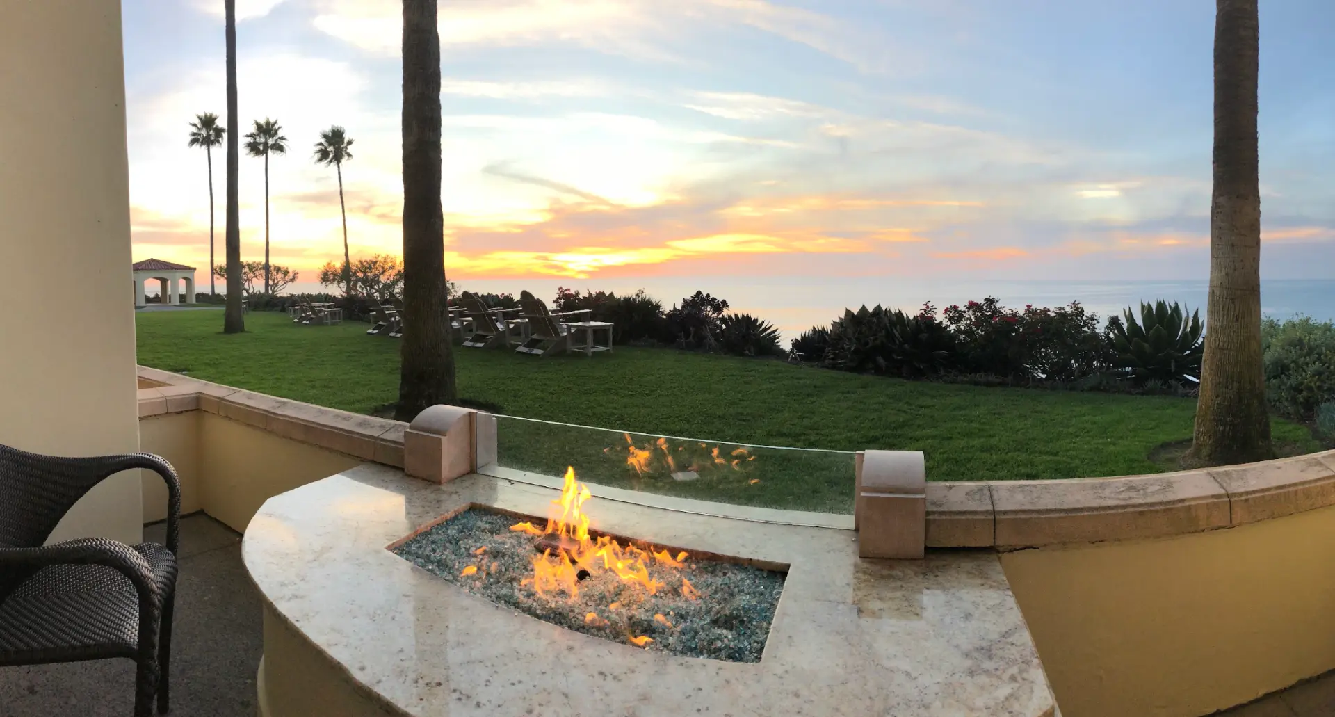 Ocean sunset seen from a patio fire pit, with palm trees and a green lawn leading to the water
