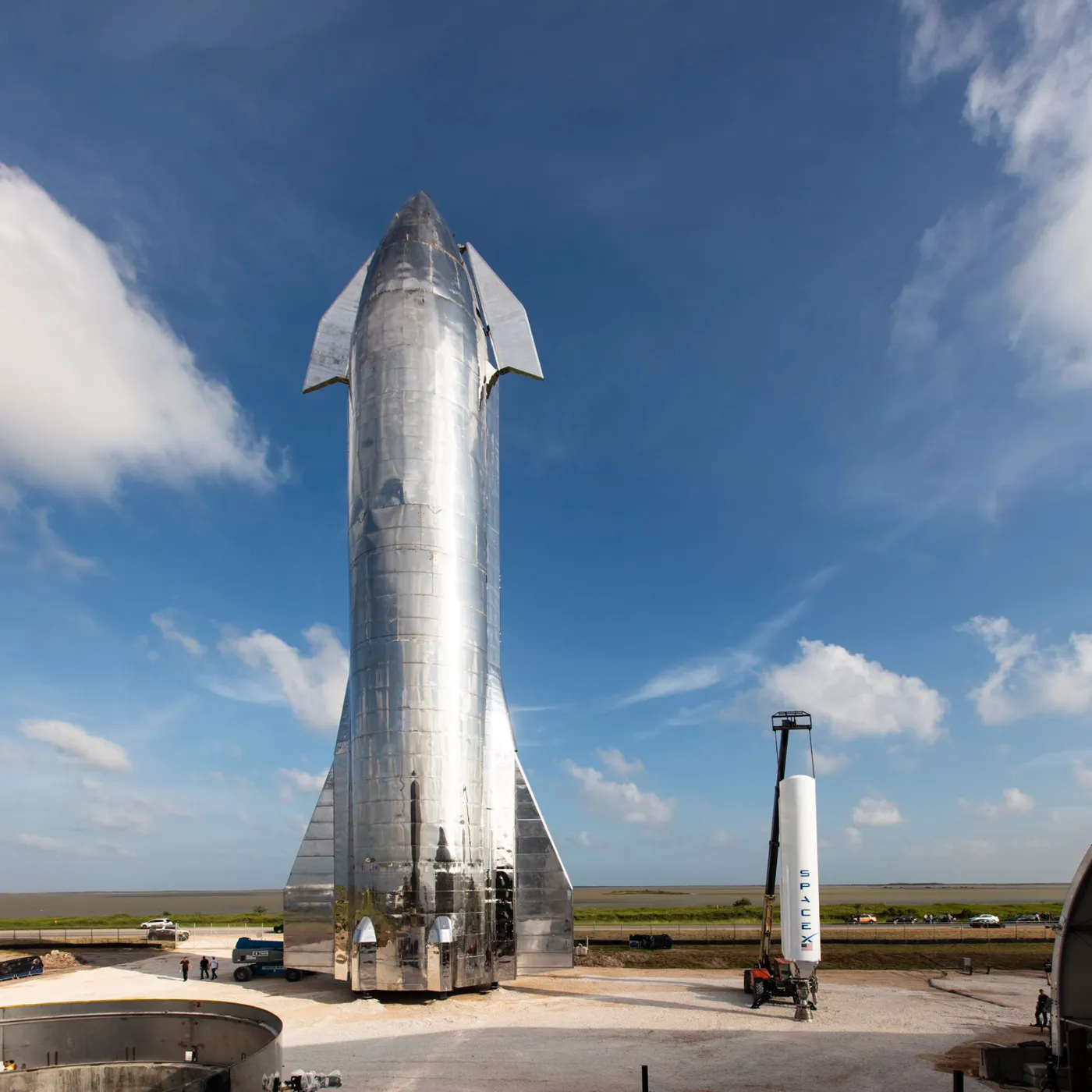 A tall stainless steel SpaceX Starship prototype standing on a gravel pad under a blue sky, next to a smaller white rocket section marked SpaceX