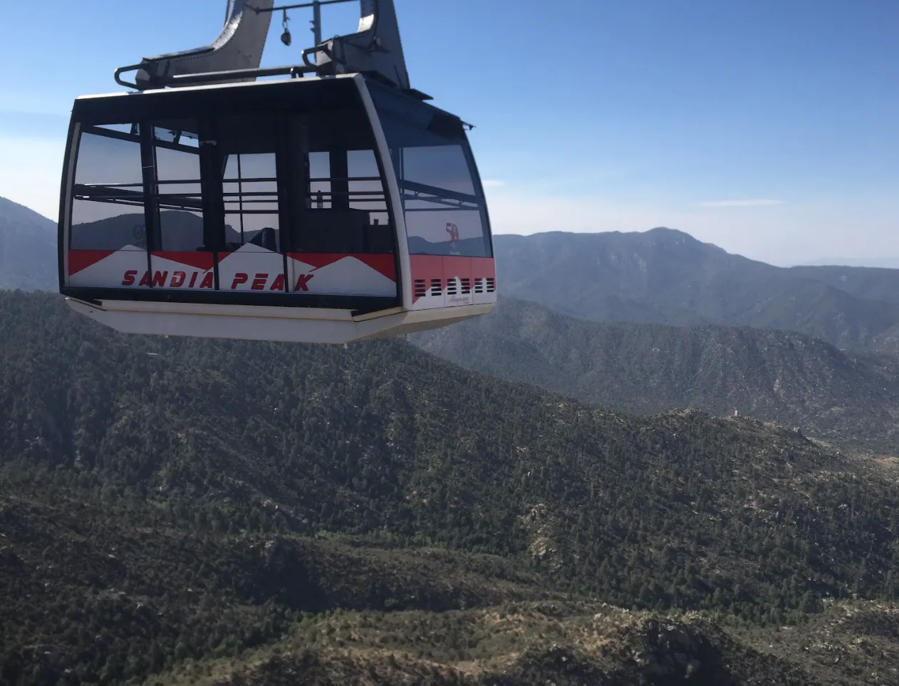 A red and white Sandia Peak Tramway car hanging over forested mountains