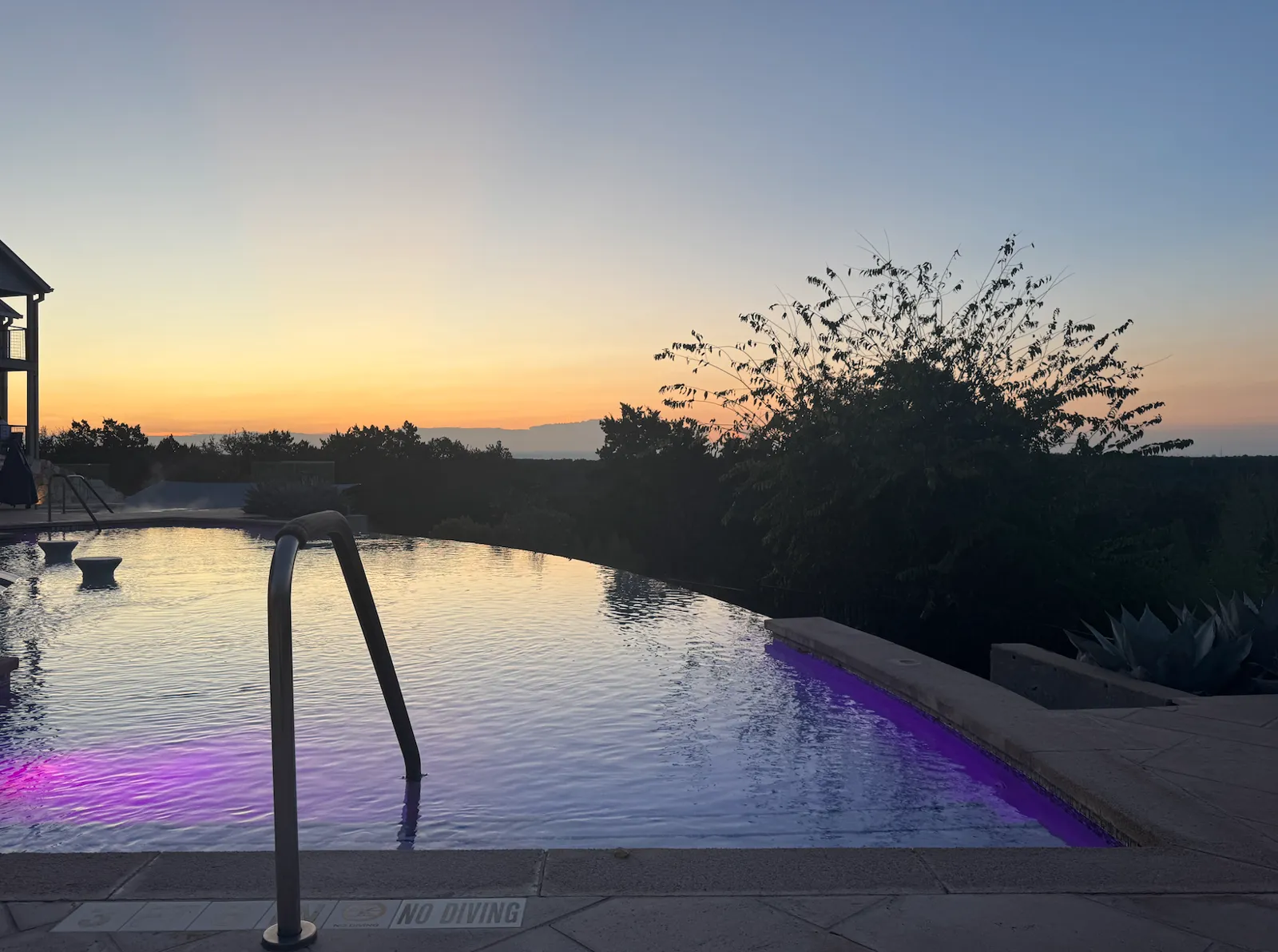 An infinity pool at sunrise with a metal handrail in the foreground, purple lights under the water and the sky turning orange over a line of trees