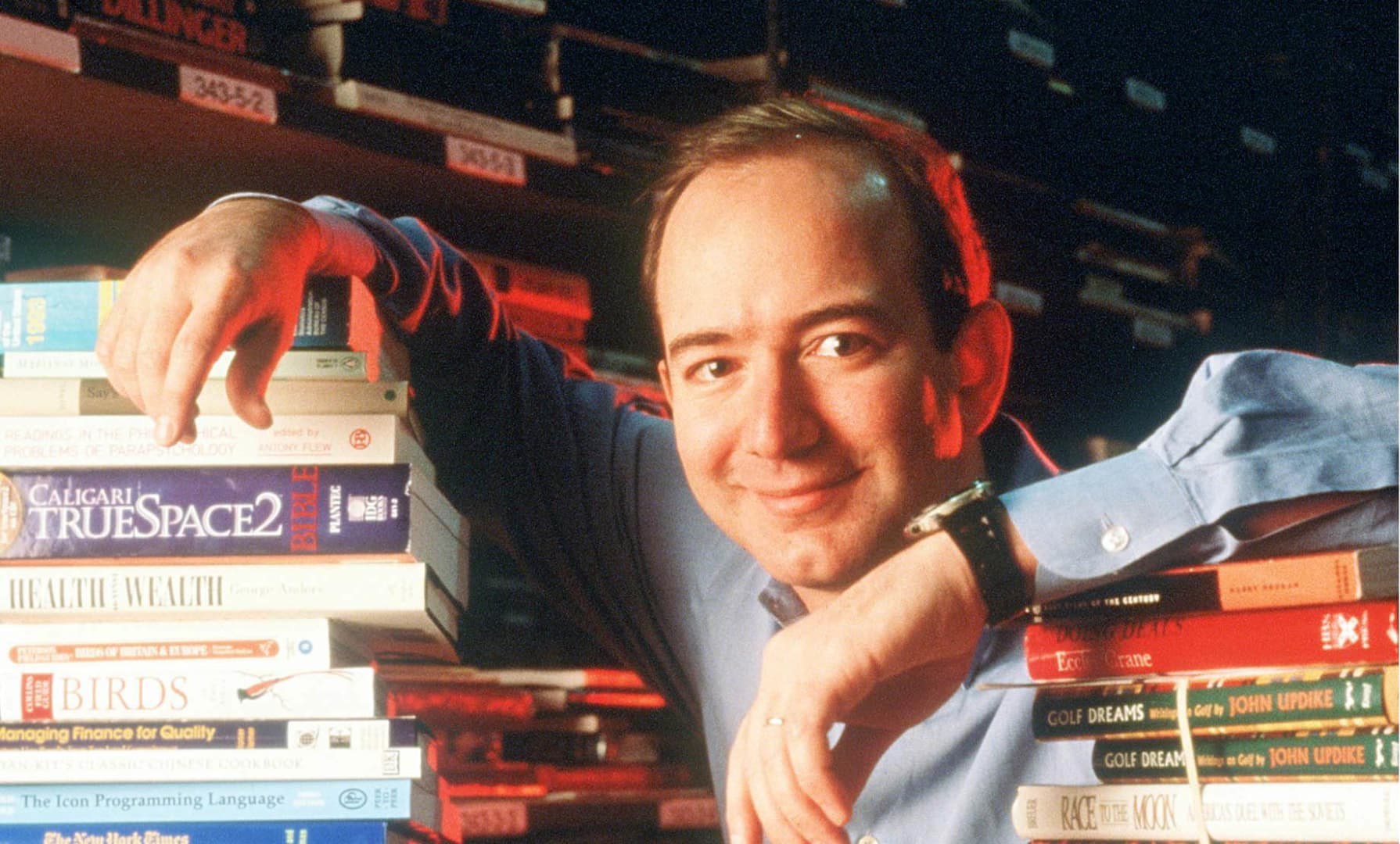 A young Jeff Bezos leaning on stacks of books in an early Amazon warehouse