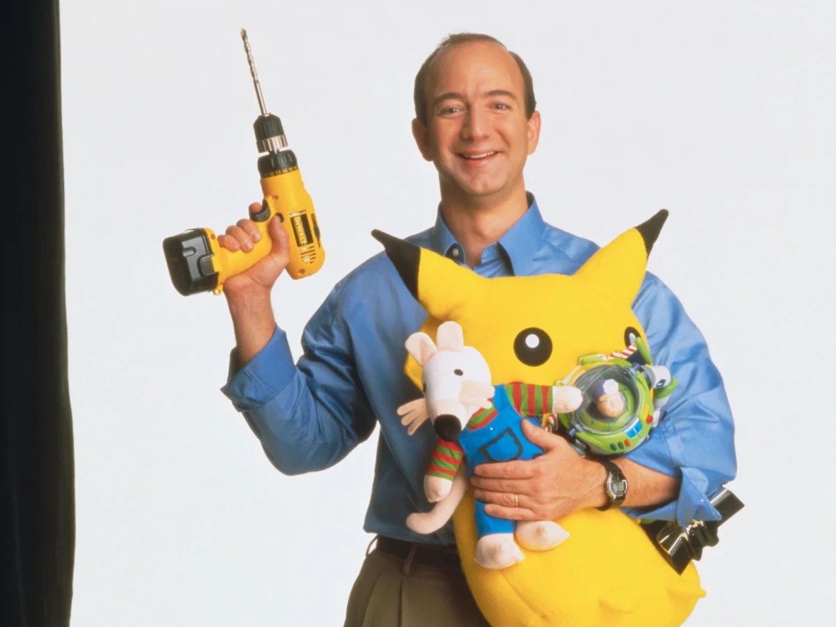 A young Jeff Bezos in a blue shirt holding a power drill and an armful of stuffed toys in the early Amazon years