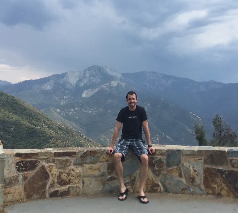 Keith Brown in a black data nerd T-shirt sitting on a stone wall at a mountain overlook under storm clouds
