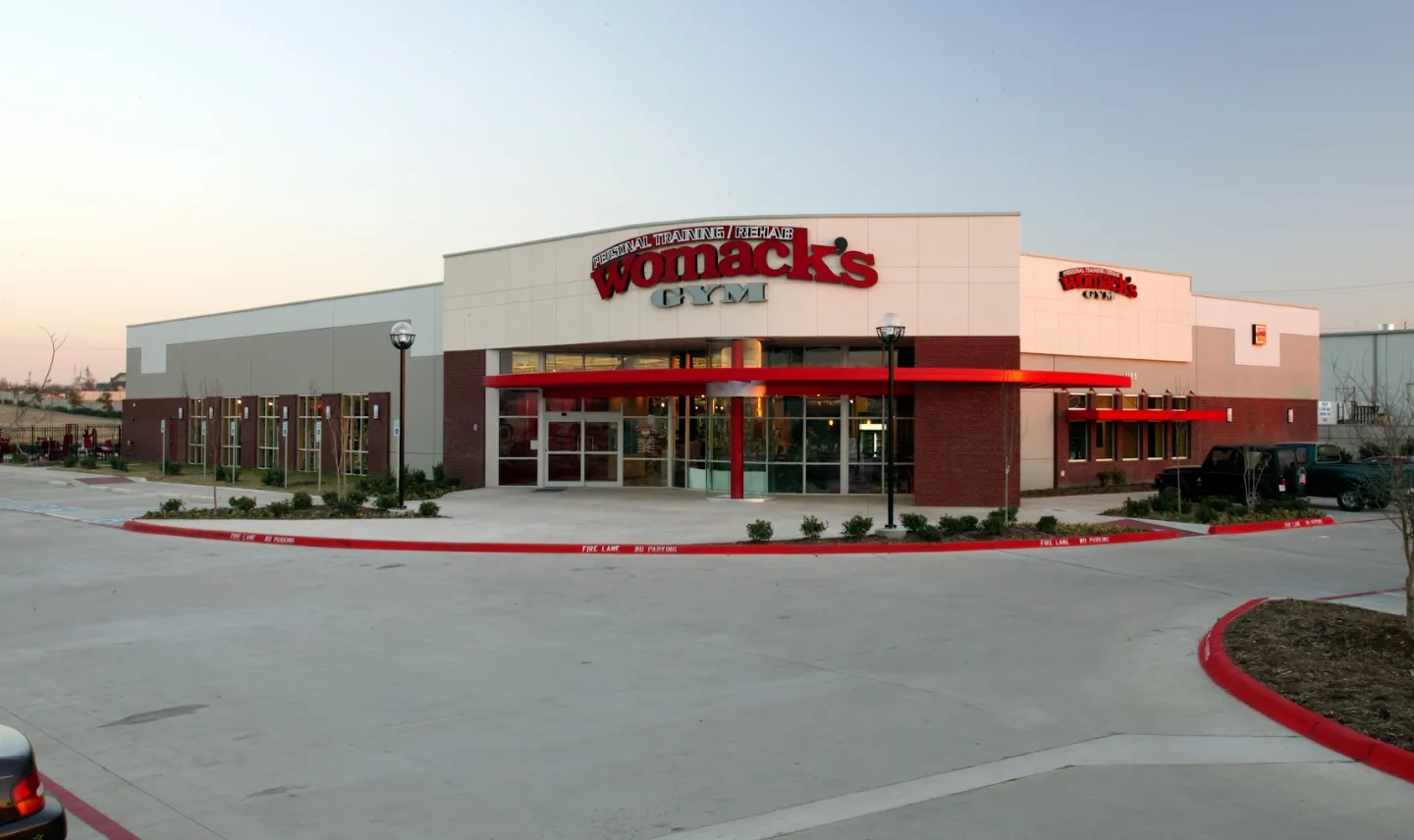 The front of Womack's Gym in Keller, Texas at dusk, a brick and stucco building with red signs reading Personal Training / Rehab and a red awning over the glass entrance