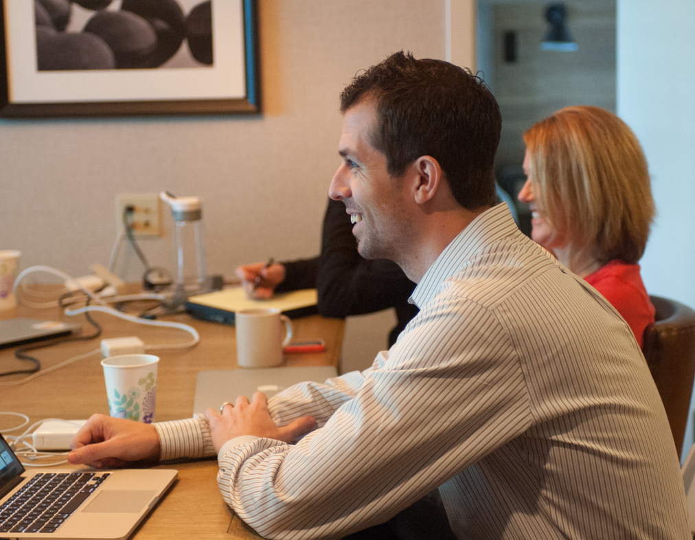 Keith Brown smiling in a team meeting at a conference table with a laptop, two colleagues beside him