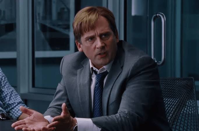Steve Carell as Mark Baum in The Big Short, sitting at a conference table and gesturing while making a point