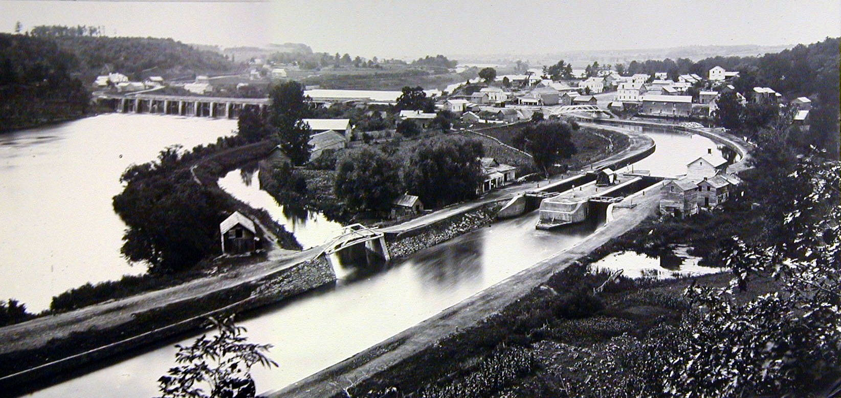 Black and white photo of the early Erie Canal and its stone aqueduct crossing the Mohawk River at Rexford, New York, with a town and locks along the water