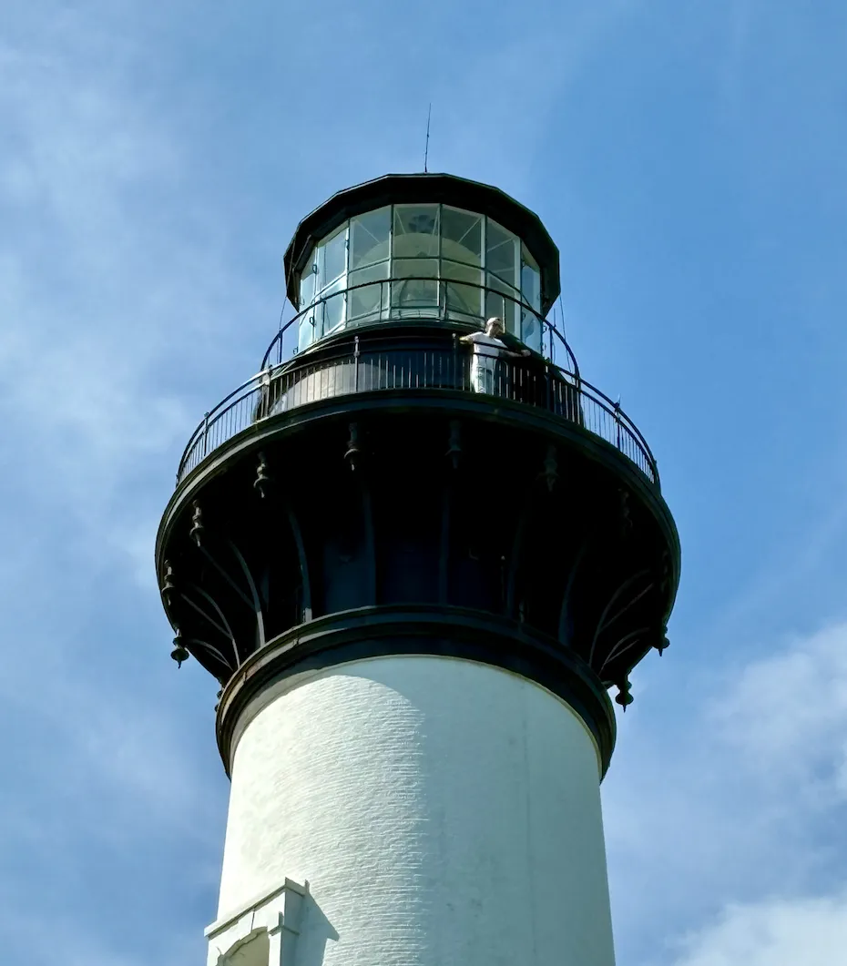 A white lighthouse with a black iron lantern room against a blue sky, with Keith Brown standing on the gallery railing near the top