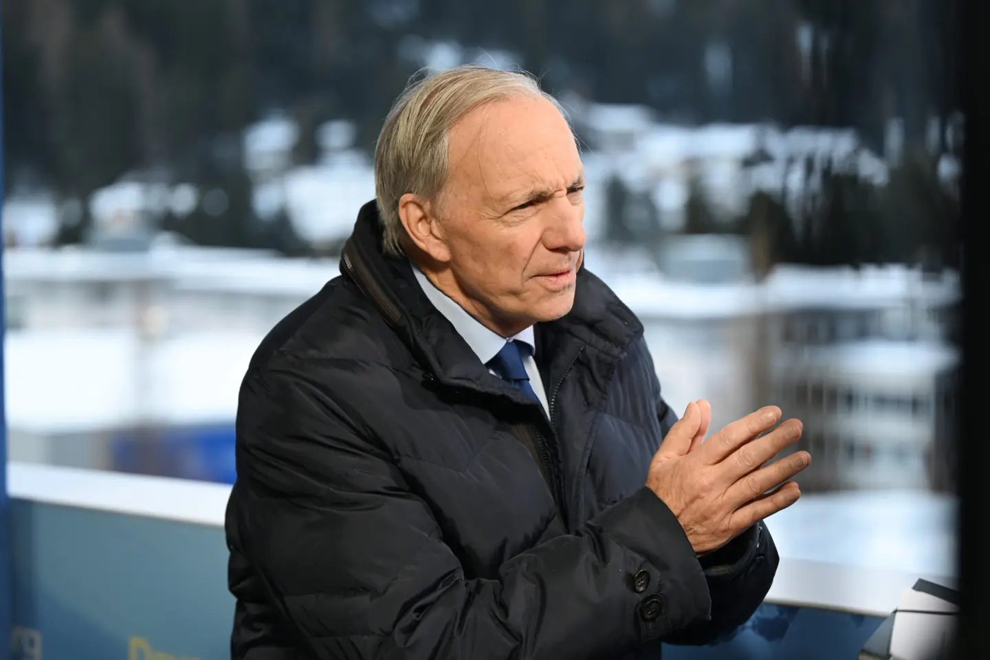 Ray Dalio in a dark winter coat and blue tie, speaking during a television interview with snowy Davos buildings behind him