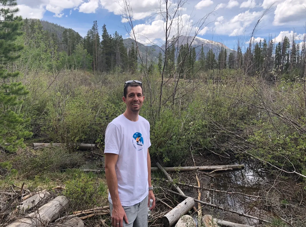 Keith Brown smiling on a trail in the Colorado mountains, with pine trees and a snow-capped peak behind him