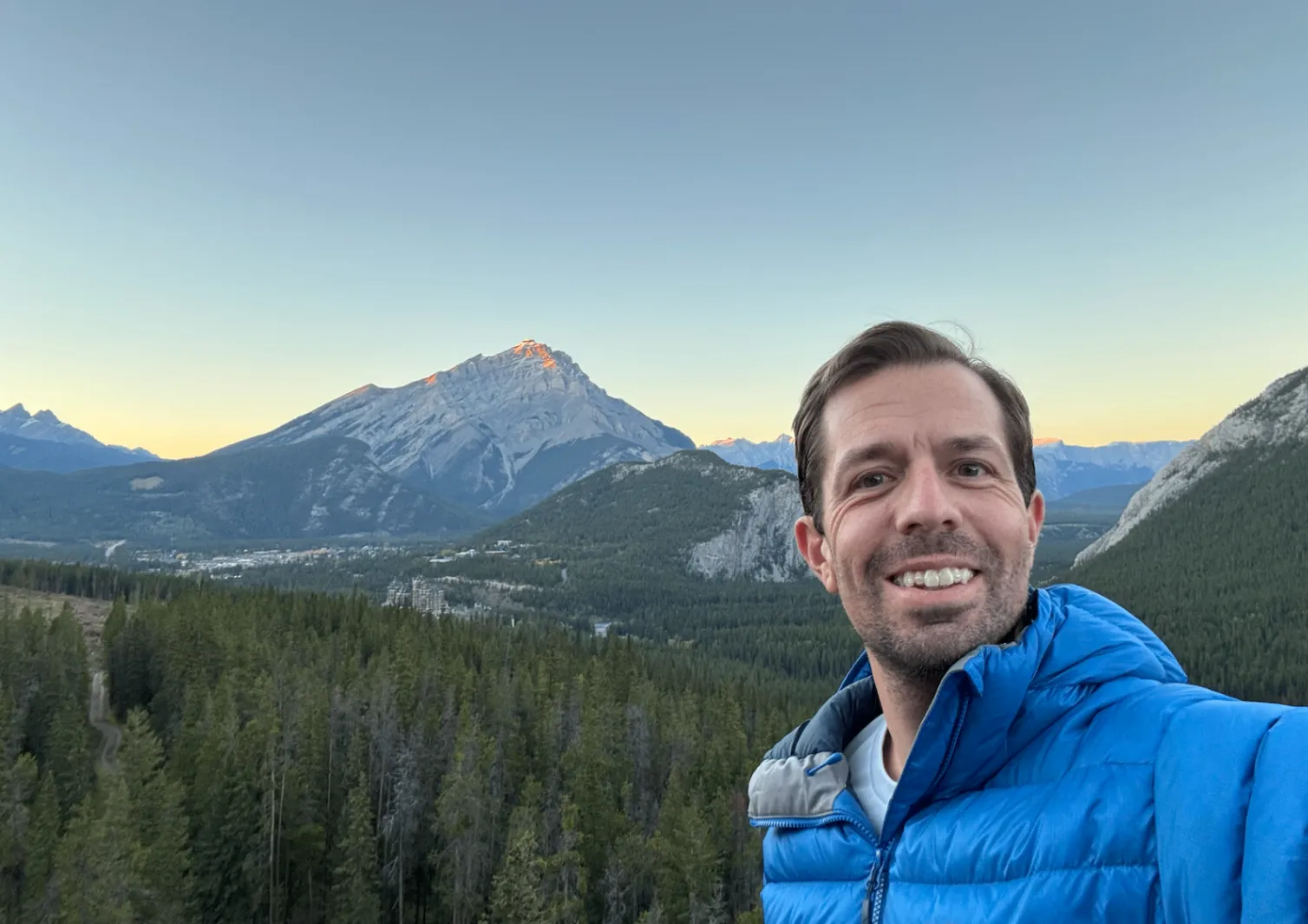 Keith Brown in a blue jacket smiling at sunrise above Banff, Alberta, with Cascade Mountain and the town in the valley behind him