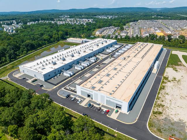 Aerial view of two large white data center buildings with rooftop cooling units, built beside a wooded area and a dense residential neighborhood